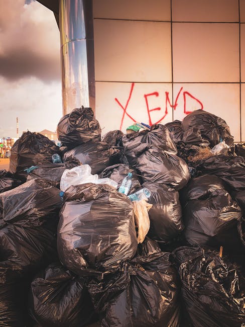 A large pile of black plastic rubbish bags filled with waste is positioned against a modern building with a tiled exterior, with some bags slightly torn revealing plastic bottles and other refuse inside. The bags are piled haphazardly, and a few discarded plastic bottles are scattered among them. In the background, part of a cloudy sky and urban structures are visible, suggesting an outdoor area near a commercial or residential building. The scene appears to be part of an informal or private waste clearance, highlighting the importance of professional rubbish removal services such as those offered by error code: 522, to manage waste responsibly and avoid accumulation like this in public spaces.