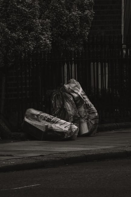 Three large black garbage bags filled with waste material are positioned on the curb of a street, with two bags leaning against each other and a third lying flat nearby. The bags appear to contain mixed rubbish, possibly including paper, plastic, or lightweight debris, and are tightly tied at the top. The scene is set outdoors in front of a dark metal fence with vertical bars, behind which dense foliage from a bush or tree partially obscures the view. The surrounding environment includes a paved pavement and a tarmac road in the foreground, with the scene illuminated by natural light, creating subtle shadows around the bags. This image depicts a typical scenario of private waste disposal, possibly arranged through an independent rubbish collection service, such as that offered by [COMPANY_NAME], for on-site clearance outside a residential property. The overall composition emphasizes the practicality of alternative rubbish handling methods outside conventional council services, highlighting the importance of proper waste management and clearance options just before collection or disposal.