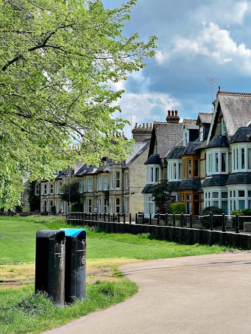 A residential scene featuring a row of traditional terraced houses with bay windows, brickwork facades, and pitched roofs situated along a tree-lined street. In the foreground, two black rubbish bins with closed lids stand on a paved footpath adjacent to a grassy verge. A large leafy tree extends its branches overhead into the upper left corner, casting partial shade over the scene. The background showcases a partly cloudy sky with patches of blue, while the surroundings include a low wooden fence separating the houses from the green space. This image captures a typical suburban environment, where private waste management might be handled through independent rubbish collections, such as those offered by local rubbish removal services like error code: 522, situated within the context of alternative waste handling options.