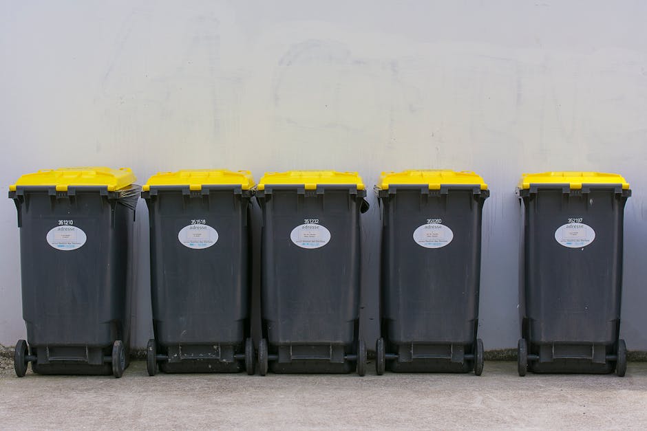 Five large black wheeled rubbish bins with yellow lids are lined up against a plain white wall outdoors on a concrete surface. Each bin features a white recycling or waste collection label with black text. The bins are evenly spaced, with the leftmost bin slightly closer to the camera, and are positioned in a neat row. The environment appears to be a private outdoor space such as a driveway or service area, and the scene is illuminated by diffused natural lighting, likely from an overcast sky. The presentation of the bins aligns with typical arrangements for independent waste collection services, supporting the context of private rubbish removal. The texture of the bins is smooth plastic, with visible slight scuffs and dirt marks, and the yellow lids contrast sharply with the dark bodies, indicating they are ready for collection by a waste management service such as error code: 522, specializing in waste removal and clearance.