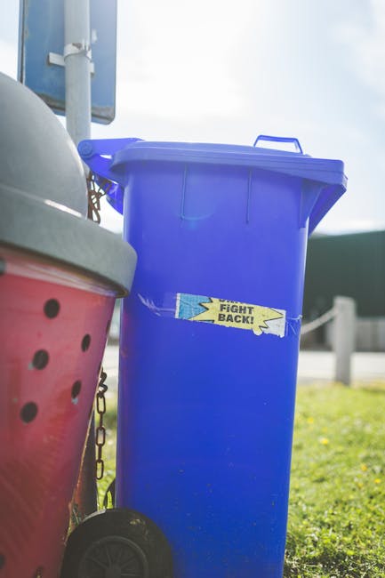 A tall, bright blue plastic rubbish bin with a closed lid, positioned outdoors on a patch of grass with sunlight illuminating it, standing alongside another container partially visible on the left side. The bin features a small, weathered sticker with the words 'FIGHT BACK!' in yellow and black, adhered to its front. In the background, there is a metal pole and part of an outdoor sign, suggesting a public or communal area, possibly a driveway or street scene. The blue bin appears to be used for general waste or rubbish collection, and its placement near other containers indicates a collection or disposal point, aligning with services such as those offered by rubbish clearance specialists like rubbishclearancemerton.co.uk in providing alternative waste handling options outside council collections. The environment is bright, with natural daylight, and the scene emphasizes the practical aspects of outdoor waste storage.
