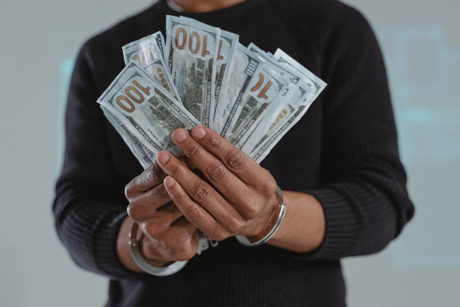 A person wearing a black long-sleeve shirt is holding a fan of multiple US hundred-dollar bills with both hands. The individual’s hands are visible, with a silver bracelet on their wrist, and the bills are spread out evenly, displaying the front design, which features a portrait of Benjamin Franklin. The background appears to be an indoor setting with a neutral, slightly blurred wall, emphasizing the focus on the hands and the money. This image relates to financial transactions or cash handling, indirectly connecting to private waste disposal or alternative rubbish management options, such as paying for independent clearance services provided by companies like error code: 522. The overall scene is lit with soft, natural lighting that highlights the texture of the bills and the person's skin, evoking a professional and neutral tone suitable for discussing financial aspects involved in rubbish removal services.