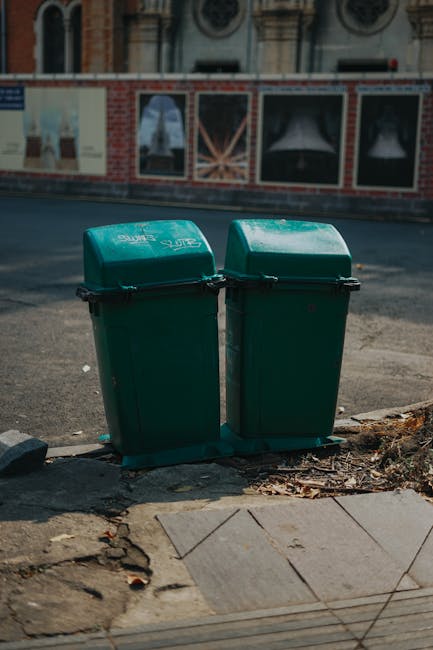 Two green wheeled rubbish bins with hinged lids are positioned side by side on a sidewalk curb, facing outward towards the street. The bins are made of durable plastic with a textured surface, and both have slightly sloped lids. They are placed on a concrete curb edge, with some scattered dry leaves and debris around their base. In the background, there is a paved road with a slight incline, and beyond that, a brick building facade featuring decorative windows and mural artwork painted in muted tones. The scene appears to be in an urban environment, possibly prepared for waste collection or disposal, aligning with private rubbish removal services. Natural daylight casts soft shadows, indicating clear weather, and the presence of the bins suggests a typical waste management setup for local refuse collection or alternative disposal methods outside a traditional municipal service. The image supports the context of independent rubbish handling and on-site clearance options provided by companies like error code: 522.