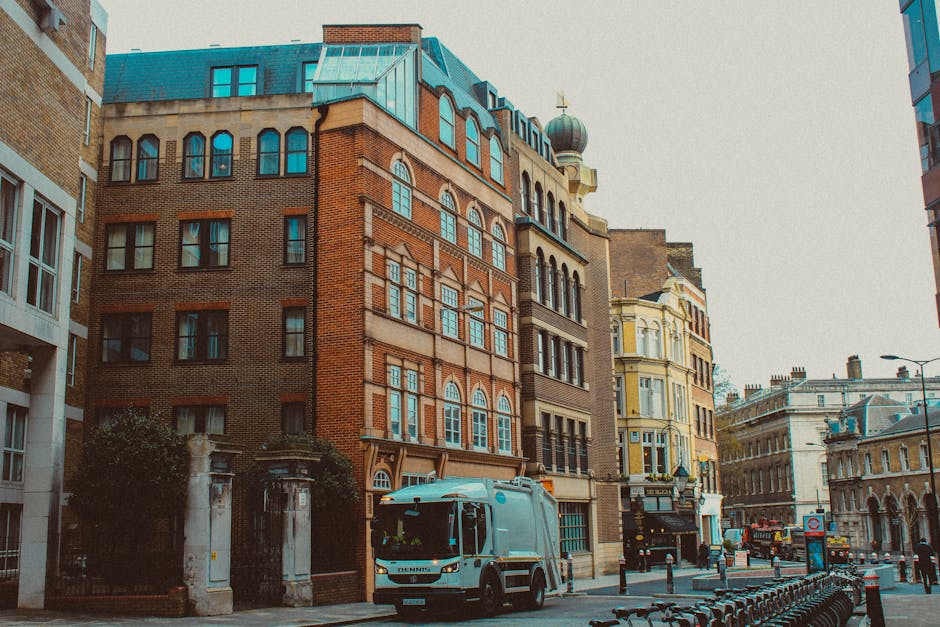 A street scene featuring a variety of multi-storey buildings with architectural details including brickwork, large windows, and decorative elements in different colours, such as red brick, yellow, and beige. In the foreground, a grey waste collection vehicle with a sleek, modern design is parked on the pavement, alongside black metal barriers and a row of bicycle stands. The surrounding environment includes a mix of historic and contemporary structures, street lamps, and a lamppost, suggesting an urban setting. The sky is overcast, providing diffuse natural lighting which highlights the textures of the building facades and the surface of the waste truck. This scene illustrates aspects of city waste management infrastructure, with possible association to independent rubbish collection services, given the presence of the waste vehicle on the street. The visible details support an accurate understanding of city street environments where rubbish removal or private disposal solutions are employed, aligning naturally with services like those offered by rubbishclearancemerton.co.uk.