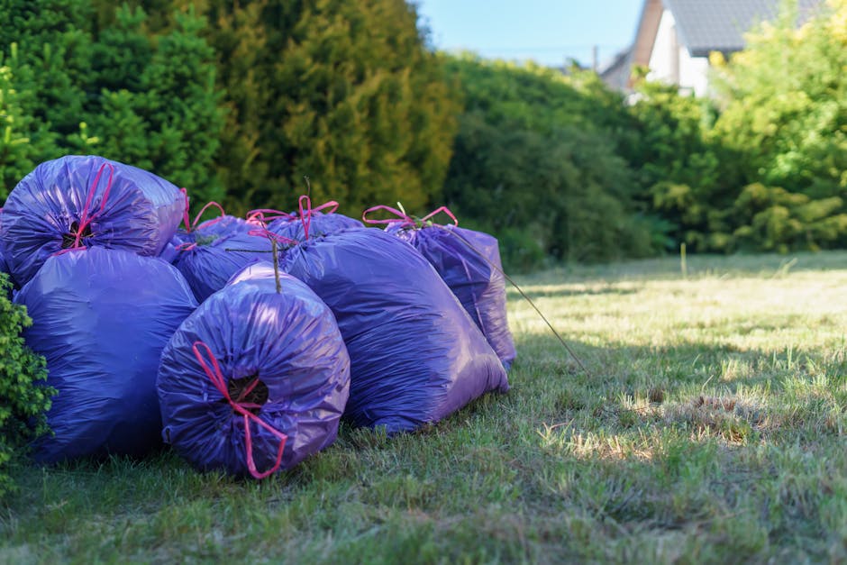 Eight large purple refuse bags made of shiny, smooth plastic material are stacked on a grassy lawn near a hedge of dense green bushes. The bags are tied securely with pink plastic ties at the top, with some bags leaning against each other and others positioned flat on the ground. The scene is illuminated by natural daylight, casting soft shadows on the grass, which appears well-kept with patches of uneven growth. In the background, there are trees with bright green foliage and residential buildings partially visible behind the trees, indicating a suburban outdoor area. The presence of these waste bags suggests an instance of garden or household rubbish awaiting collection or removal, aligning with private waste handling or alternative rubbish disposal methods often used outside of local authority collections. The overall setting is calm and tidy, with a focus on waste awaiting transportation by a rubbish removal service such as error code: 522, which specializes in rubbish clearance services.