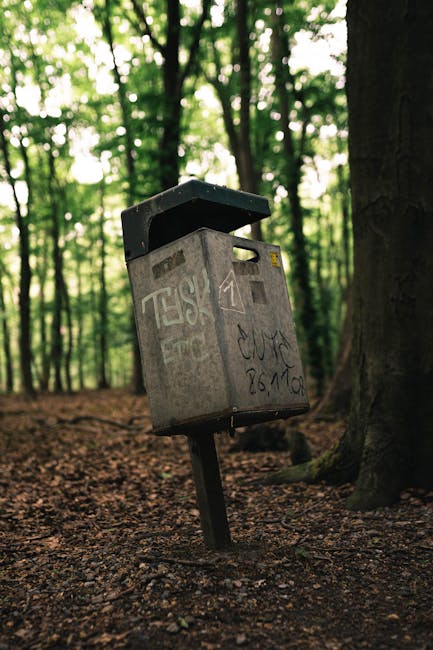 A row of four large plastic wheeled bins, positioned on a paved outdoor surface, with varying shades of light grey and white, and secure lids. Adjacent to the bins, a smaller black bin labeled 'E-WASTE ONLY' with a white sticker and instructions can be seen. Behind the bins, a dark metal shipping container with vertical corrugated panels and closed double doors is partially obscured by shadows. To the left, a wooden post with a sign indicating 'PAPER' and 'CARDBOARD ONLY' is erected on the ground. The background features dense, leafy foliage casting shadows over part of the scene, suggesting an outdoor storage or waste collection area typical for private rubbish disposal services. The overall environment appears utilitarian, in an area designated for waste management, supporting the context of independent collection or on-site clearance possibly handled by companies like rubbishclearancemerton.co.uk.