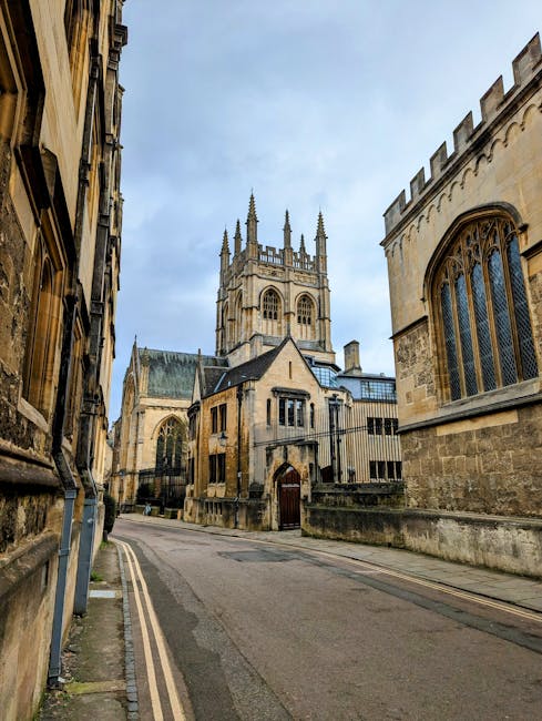 A narrow, slightly curved street flanked by historic stone buildings with a large medieval-style church in the background featuring a prominent square tower with multiple spires and Gothic architectural details. The buildings on both sides have weathered stone facades, arched windows with intricate tracery, and leaded glass panels. The street surface appears to be asphalt, with a small curb and a narrow sidewalk on the left side. The sky overhead is cloudy, casting diffuse light over the scene, which reflects an older urban environment. This setting may relate to private or independent waste disposal, as the scene shows an area unlikely to have visible waste collection vehicles or infrastructure, and the historical context suggests it might be part of a location where rubbish clearance services are sometimes sought for property clearance or renovation projects. The image is taken from a low vantage point, emphasizing the height of the church and surrounding architecture, with no visible activity or vehicles.
