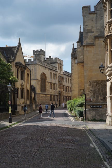 A narrow street in an historic town features a cobblestone surface with a slight incline, bordered by old stone buildings with ornate architectural details and large windows. On the left, a church or cathedral with pointed gothic features, arched windows, and battlemented tower rises above the street. Several pedestrians, dressed in casual clothing, are seen walking along the pavement on both sides of the road, some near the base of the church, while others are further ahead. To the right, a stone wall with a corner feature and a sign indicating an address or street name is partially visible, with lush green bushes alongside the wall. The sky above is mostly cloudy with patches of blue, casting diffuse natural light over the scene. The environment appears quiet and clean, reflecting an area that may be part of a historic or tourist district where private or independent rubbish removal services, such as those offered by rubbishclearancemerton.co.uk, could be utilized for upkeep and waste management.