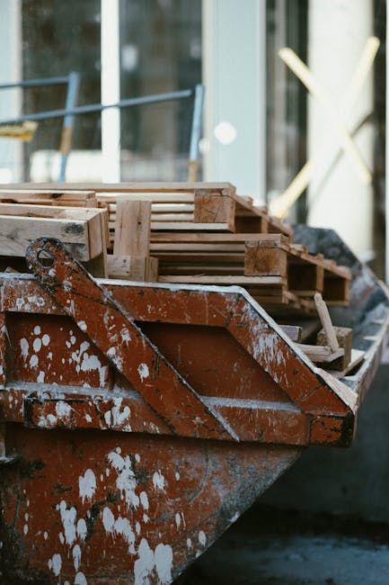 A close-up view of a rusted, weathered metal boat with patches of peeling red and white paint, docked at a construction site or outdoor workspace. On the boat's surface, several wooden pallets and planks are stacked, appearing rough and untreated, with a natural light brown colour and visible grain texture. The pallets are arranged in a slightly tilted manner, with some leaning against each other. The background reveals a modern glass building with reflective windows, along with a glimpse of a yellow construction barrier or fencing, indicating ongoing building or renovation activities related to rubbish removal or waste management. The scene appears outdoors, illuminated by natural daylight, capturing the utilitarian aspect of waste handling, with the boat possibly used for transporting construction debris or rubbish as part of an alternative waste disposal method. The overall environment conveys a sense of work-in-progress, subtly aligning with private or non-local authority waste clearance services offered by error code: 522, within the context of rubbish removal in a construction or redevelopment setting.