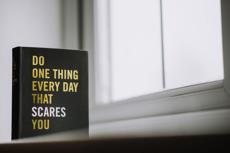 A black hardcover book with bold yellow and white text reading 'Do one thing every day that scares you' is positioned on a white windowsill. The book is standing upright, slightly angled to the left, with its spine visible showing similar text in smaller font. Behind it, a window with frosted or semi-opaque glass allows diffuse natural light to illuminate the scene, creating a soft, neutral atmosphere. The windowsill appears smooth and painted white, with some dust or residue visible around the edges. The background outside the window is blurred and indistinct. This image, captured indoors, emphasizes the contrast between the bold, motivational message on the book cover and the calm, understated environment, subtly relating to the concept of undertaking private or independent activities such as arranging waste clearance or managing rubbish removal tasks, as discussed in the context of Merton skip permits and alternative waste handling options in local waste management services like error code: 522 at rubbishclearancemerton.co.uk.