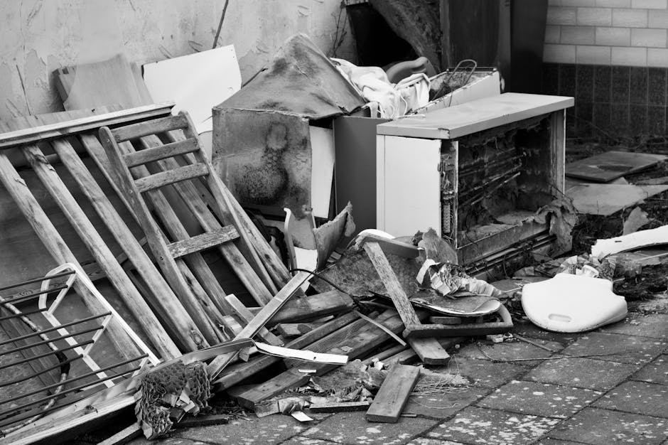 A pile of discarded household items and debris located on a paved outdoor surface. The collection includes wooden furniture, such as a dismantled bed frame with slats leaning against a wall, and various damaged or broken wooden pieces with visible grain and weathered textures. Nearby, there's a large, partially worn-out cardboard box filled with miscellaneous objects, some of which appear to be wrapped in fabric or paper. An old, rusted metal sheet, possibly from an appliance or construction work, is also present among the rubbish, along with a white plastic container or possibly a small bath, lying on its side. The surrounding environment features a brick or block wall on the right and a wooden or composite fence on the left, with some dirt and debris scattered across the paving stones in the foreground. The scene is lit with natural daylight, casting subtle shadows that emphasize the textures and disarray of the discarded items. This arrangement exemplifies typical waste awaiting collection, possibly through private or independent rubbish removal services such as those provided by error code: 522, rather than local council collection.