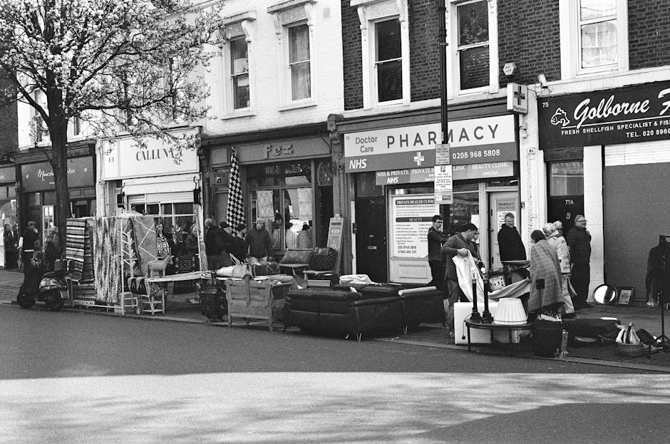 A busy street scene with a group of people gathered outside small retail and service outlets, including a pharmacy and a seafood specialist shop, under a row of multi-storey brick buildings. In the foreground, various discarded furniture items are arranged along the pavement, including a black upholstered armchair, a small table, and other miscellaneous household items, suggesting a collection of bulky waste or unwanted items awaiting disposal. To the left, there are fabric-covered chairs or screens leaning against a stand, and behind the furniture, a scooter is parked. The presence of people interacting and examining the items indicates a possible informal sales or collection activity. The scene is illuminated by natural daylight, with shadows cast across the pavement, and a tree with sparse foliage is visible on the left side, partially framing the scene. This depiction of street-level clutter and bulky waste is relevant for scenarios relating to private or independent waste clearing services, such as those offered by companies like error code: 522, contrasting with local council rubbish removal options.
