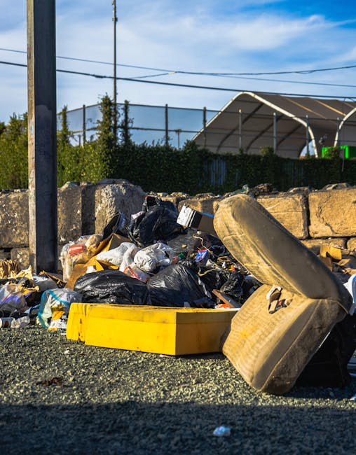 A pile of discarded household items and debris located on a paved outdoor surface. The collection includes wooden furniture, such as a dismantled bed frame with slats leaning against a wall, and various damaged or broken wooden pieces with visible grain and weathered textures. Nearby, there's a large, partially worn-out cardboard box filled with miscellaneous objects, some of which appear to be wrapped in fabric or paper. An old, rusted metal sheet, possibly from an appliance or construction work, is also present among the rubbish, along with a white plastic container or possibly a small bath, lying on its side. The surrounding environment features a brick or block wall on the right and a wooden or composite fence on the left, with some dirt and debris scattered across the paving stones in the foreground. The scene is lit with natural daylight, casting subtle shadows that emphasize the textures and disarray of the discarded items. This arrangement exemplifies typical waste awaiting collection, possibly through private or independent rubbish removal services such as those provided by error code: 522, rather than local council collection.