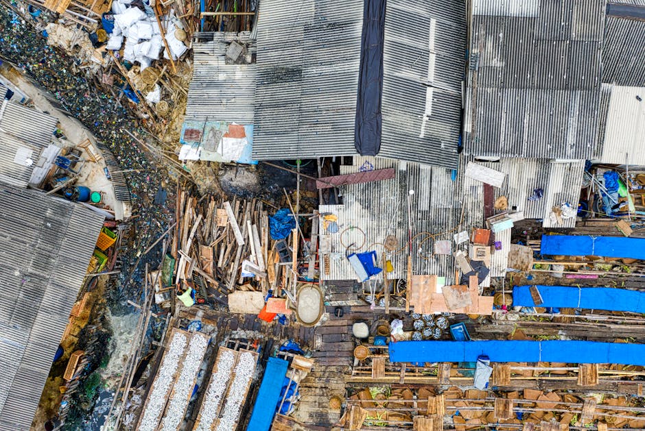 An aerial view of a cluttered outdoor area featuring a mix of construction debris, wooden planks, plastic containers, and discarded household items. The scene includes corrugated metal roofing sheets, some stacked and others scattered, with visible rust and weathered textures. To the left, there is a patch of dark, muddy ground with a pile of mixed rubbish, including plastic bottles, fabric scraps, and miscellaneous waste. Several blue tarpaulins are stretched over some sections, possibly covering stored materials. Wooden pallets and broken furniture parts are visible among the refuse, alongside plastic drums and containers of varying sizes. The environment suggests a non-organized outdoor storage or waste accumulation site, with the materials arranged haphazardly amidst structural elements, indicative of a site likely associated with alternative waste handling or private rubbish collection. The lighting appears natural, with shadows cast across the scene, offering a clear view of the diverse textures and materials present. This scene aligns with services offered by companies like rubbishclearancemerton.co.uk specialising in private rubbish removal or on-site clearance operations.